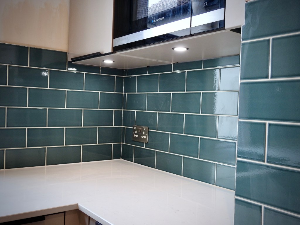 Kitchen units with lights underneath them and green metro-style tiles on the wall. White marble work surface can just been seen. 