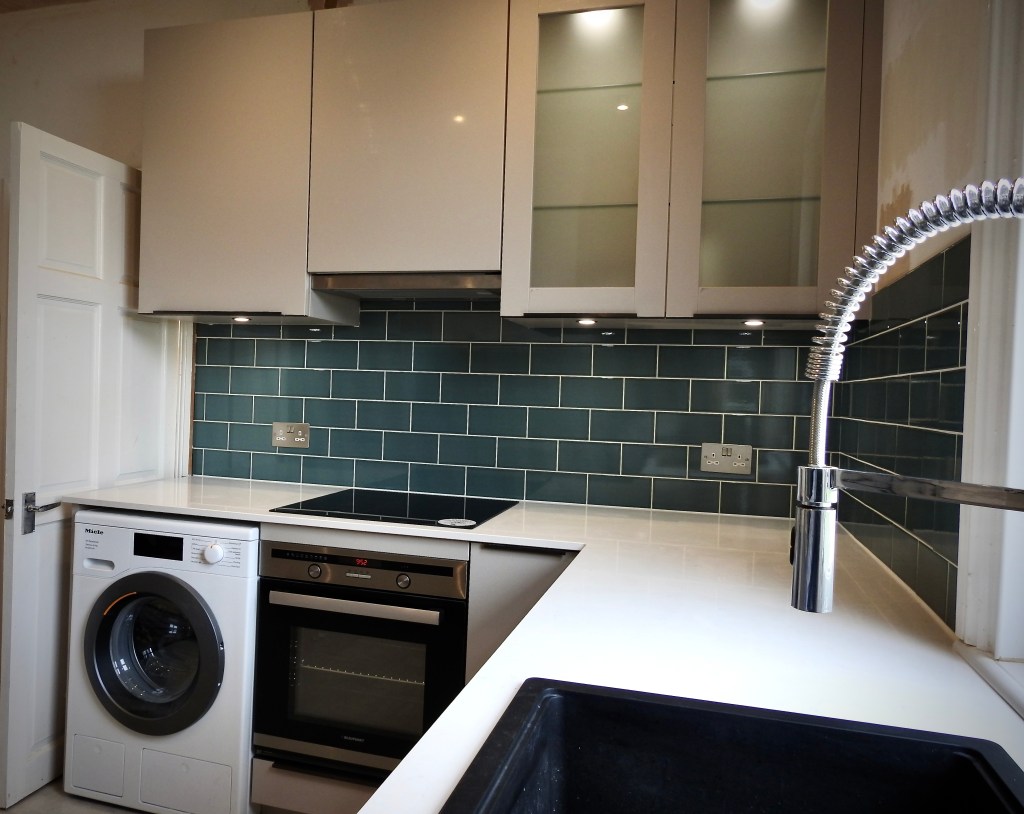 A view of part of a kitchen with the cupboards on show and the metro tiles underneath them before the white marble work surface. You can see the top of a washing machine and cooker and the kitchen tap. 