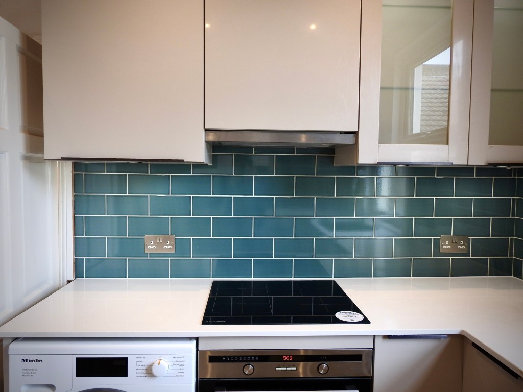 A view of part of a kitchen with the cupboards on show and the metro tiles underneath them before the white marble work surface. You can see the top of a washing machine and cooker. 